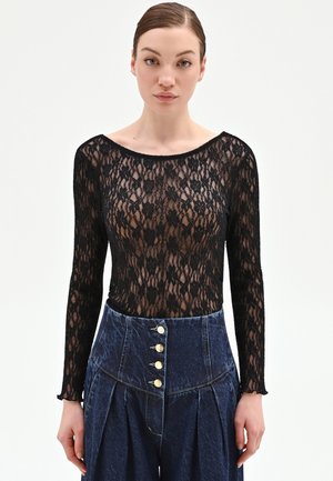 Giovane donna che indossa un top a maniche lunghe in pizzo floreale nero e pantaloni in denim blu scuro a vita alta con bottoni dorati.