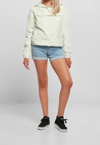 Lys grøn vindjakke med hætte, frontlomme, elastiske manchetter, parret med denim shorts og sorte sneakers.