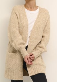 Person som bär en beige stickad cardigan över en vit skjorta och svarta byxor, med händerna löst knutna framför sig.