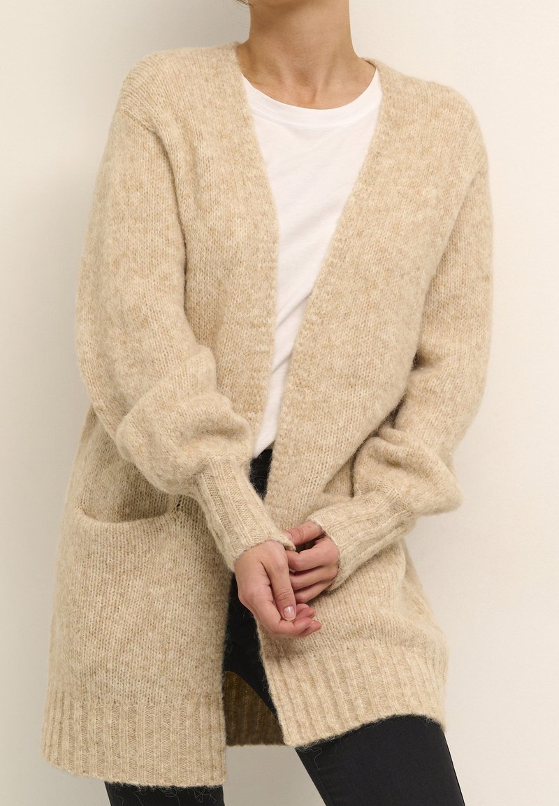 Person som bär en beige stickad cardigan över en vit skjorta och svarta byxor, med händerna löst knutna framför sig.