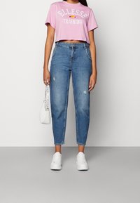 Světle růžové zkrácené tričko s bílým nápisem, zkombinované se světle modrými, opranými džíny a bílými teniskami, držící malou bílou tašku.