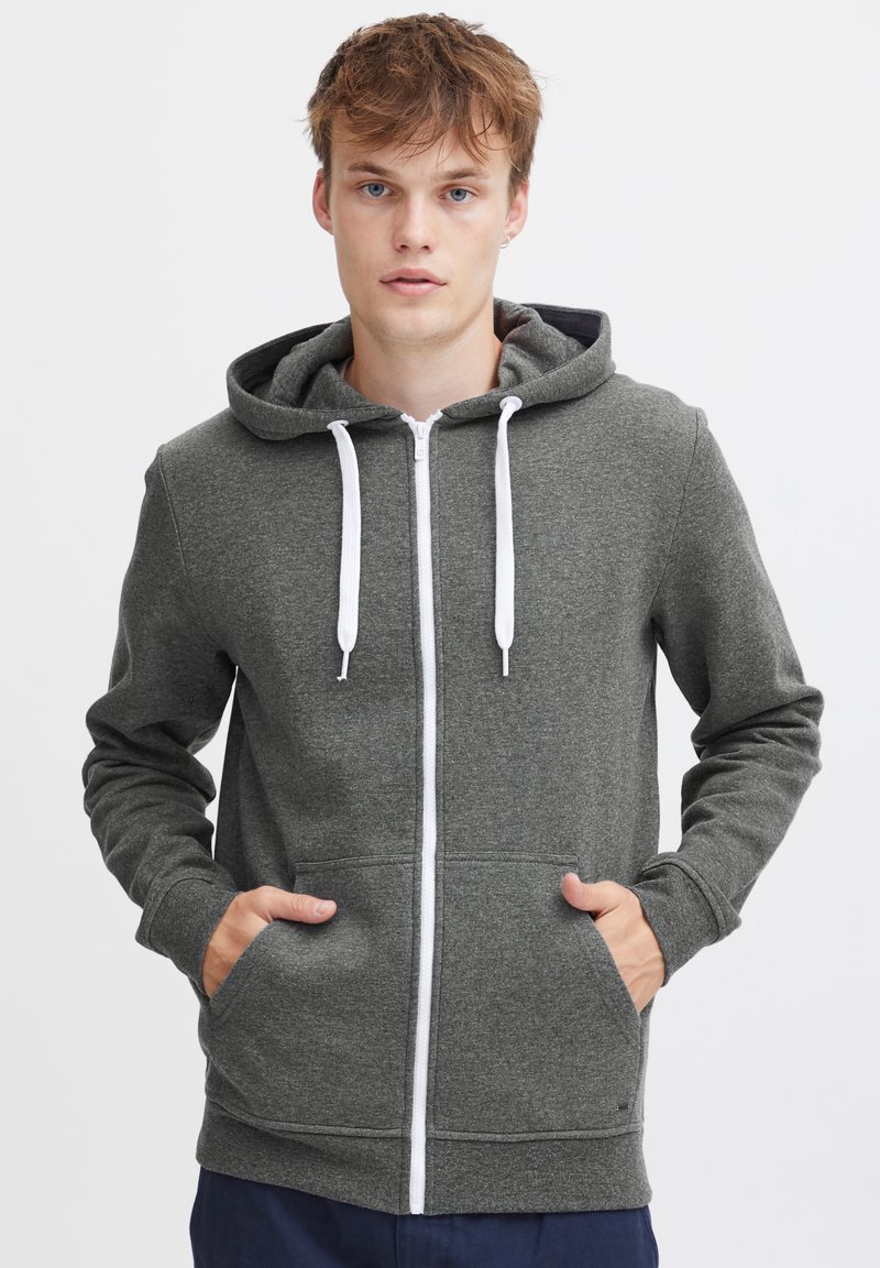 Grijze zip-up hoodie van zacht materiaal, met een trekkoordkap, voorzakken en contrasterende witte rits- en koordaccenten.