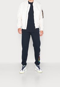 Valkoinen bomber-takki, musta T-paita, tummansiniset collegehousut monivärisellä tekstillä ja valkoiset lenkkarit vihreillä yksityiskohdilla. Silkkinen kangas, rento istuvuus.