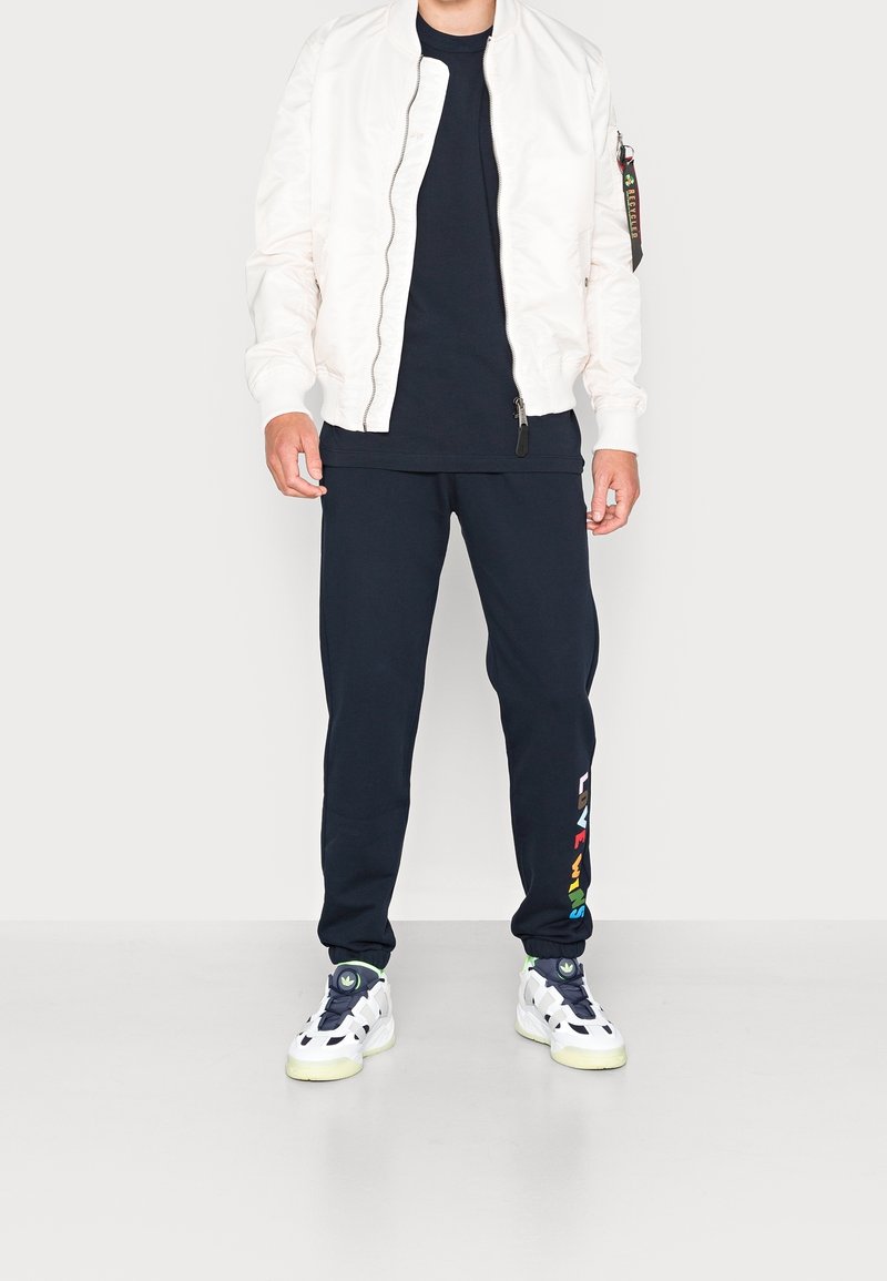 Valkoinen bomber-takki, musta T-paita, tummansiniset collegehousut monivärisellä tekstillä ja valkoiset lenkkarit vihreillä yksityiskohdilla. Silkkinen kangas, rento istuvuus.