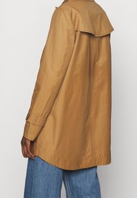 Tmavě pleti osoba nosící béžový trenčkot s buttoned rukávy přes modré džíny, zobrazená ze zadu na jednobarevném pozadí.