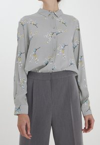 Lichtgrijs shirt met een bloempatroon in blauw, geel en wit. Heeft een knoopsluiting aan de voorkant en lange mouwen, gecombineerd met grijze broeken.