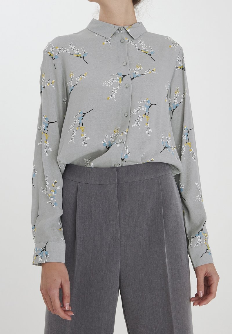 Lichtgrijs shirt met een bloempatroon in blauw, geel en wit. Heeft een knoopsluiting aan de voorkant en lange mouwen, gecombineerd met grijze broeken.