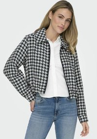 Jachetă cropped cu model houndstooth alb-negru, cu guler, închidere cu fermoar și mâneci lungi, purtată peste o cămașă albă și blugi.