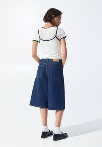 Biela tričko s čiernymi záhybmi, skombinované s širokými denimovými culottes v tmavomodrej farbe. Model má na sebe čierne topánky a biele ponožky.