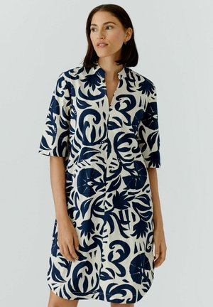 Frau trägt ein knielanges Kleid mit kurzen Ärmeln und großem schwarzem und weißem Blumenmuster, stehend vor einem schlichten hellen Hintergrund.