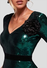 Smaragdgrøn palietkjole med dyb V-udskæring, fremhævet af en sort tekstureret blomst på skulderen og et sort fløjlsbånd i taljen.