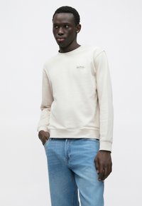 Μπεζ φούτερ με μαλακή υφή, διαθέτει στρογγυλή λαιμόκοψη, ριμπ μανσέτες και μικρό κεντητό λογότυπο στο στήθος, φορεμένο με ανοιχτόχρωμο τζιν.