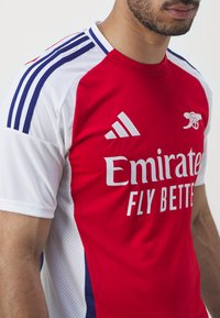 Tricou roșu și alb cu logo-ul adidas, textul "Emirates Fly Better", accente cu dungi albastre și albe pe umeri, textură respirabilă.