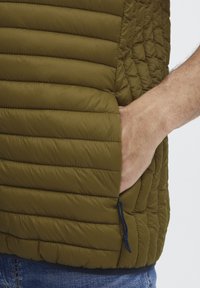 Polstret olivengrøn vest med quiltet tekstur og sidelomme med snor. Letvægtsdesign, der er velegnet til lagdeling.