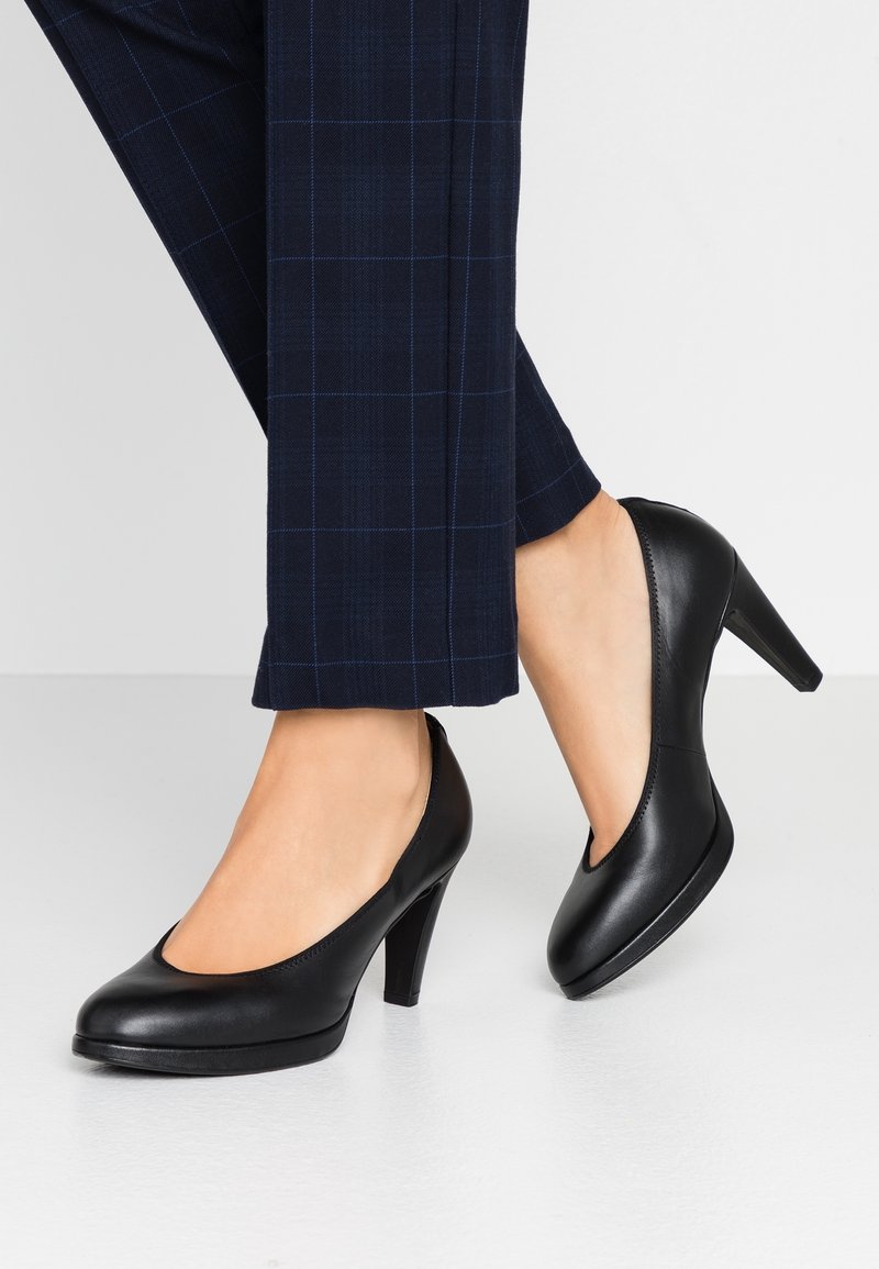 Femme portant des escarpins en cuir noir à talons moyens avec un pantalon tailleur à carreaux bleu foncé, debout sur un sol clair.