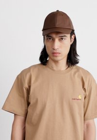 Casquette de baseball marron avec visière courbée, portée avec un t-shirt beige à manches courtes présentant un petit logo rouge et jaune sur la poitrine.