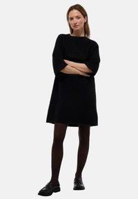 Robe en maille noire à coupe ample, avec des manches courtes et un col rond. Associée à des collants sombres et des mocassins noirs. Design simple et texturé.