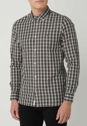 Homme portant une chemise à manches longues boutonnée avec un motif à carreaux noir, blanc et rouge et un pantalon noir, debout devant un fond uni.