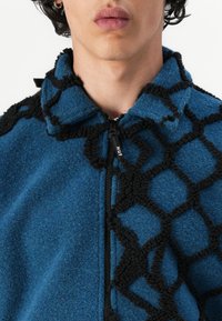 Fleece jacket in blue, featuring a textured black geometric pattern on one side, high collar, and zipper closure.