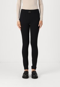 Pantalons ajustés noirs avec une texture lisse, présentant un petit détail de ceinture métallique et un design élégant, longueur cheville.