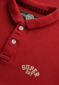 Polo rouge en tissu texturé, doté d'une patte de boutonnage avec deux boutons blancs et du logo brodé "SUPER DRY" en crème.