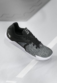 Čierna športová obuv s vrstvenou sieťovinou a pevným čiernym akcentom, s bielou podrážkou a logom Reebok na jazyku a strane.