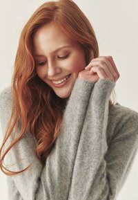 Hall hõbedase värvusega cashmere'i sviiter, millel on pikad varrukad, soonikud ja pehme, tekstureeritud pind. Mudel hoiab varrukatest kinni ja tal on lainelised punased juuksed.