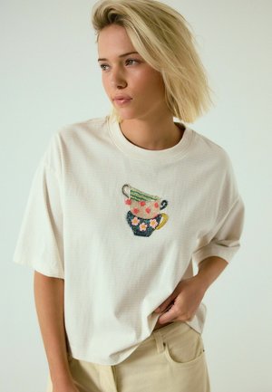 Blonde Frau trägt ein cremefarbenes, übergroßes T-Shirt mit bestickten gestapelten Teetassen, schaut zur Seite und hat die Hand in der Tasche der beigen Hose.