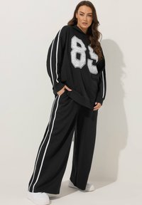 Felpa nera con cappuccio, caratterizzata da un grande numero "85" in bianco, abbinata a pantaloni neri larghi con strisce bianche laterali e tasche frontali.