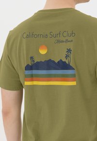 Oliivinvihreä puuvillapaita, jossa on graafinen suunnittelu vuorista, palmuista, auringosta ja monivärisistä vaaka-viivoista. Suunnittelun yläpuolella on teksti "California Surf Club Malibu Beach".