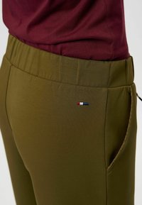 Olive krāsas sporta bikses ar elastīgu jostu, kurām pie kabatas ir neliels izšūts logo sarkanā, baltā un zilā krāsā.