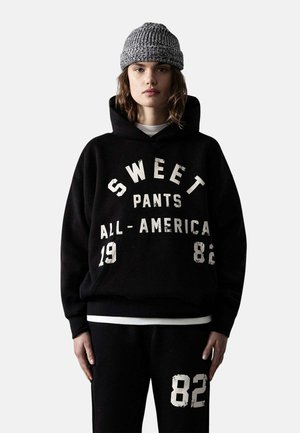 Joven mujer con una sudadera negra con capucha con texto blanco "SWEET PANTS ALL-AMERICA 1982" y pantalones deportivos negros con el número "82" en una pierna.