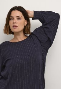 Blusa azul marino con rayas plateadas verticales, escote amplio y mangas abullonadas. Confeccionada en una tela texturizada, que proporciona un ajuste relajado.