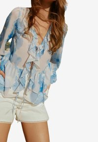 Lätt blus med blå akvarellblommönster, volang framtill och vid ärmarna, samt en knytning framtill. Matchas med vita shorts med spetsdetaljer.