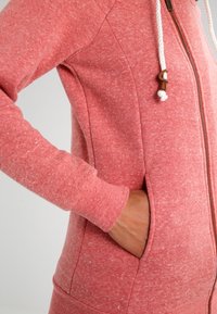 Sudadera con cremallera en tela coral jaspeada. Cuenta con cordones con detalles de madera, bolsillos laterales y puños acanalados para un ajuste ceñido.