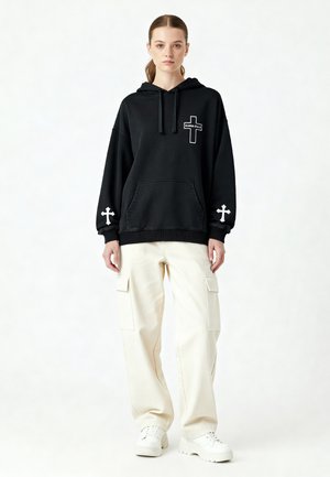 Hoodie noir surdimensionné avec poche frontale, designs de croix blancs sur les manches et la poitrine, associé à un pantalon cargo crème et des baskets blanches.
