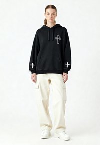 Hoodie oversize nera con tasca frontale, design a croce bianca sulle maniche e sul petto, abbinato a pantaloni cargo color crema e sneakers bianche.