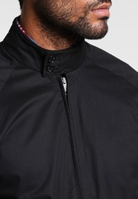 Giacca nera con colletto rialzato, chiusura con zip e due accenti a bottoni. Realizzata in tessuto leggero e liscio con una leggera lucentezza.