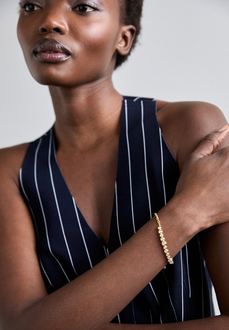 Χρυσό βραχιόλι με υφασμάτινους συνδέσμους, φορεμένο σε καφέ χέρι, σε φόντο ναυτικού και λευκού ριγέ υφάσματος.