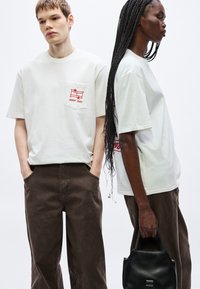 Camisetas blancas de algodón con logo rojo y detalle de bolsillo, combinadas con pantalones marrones de pierna ancha. Un modelo sostiene un bolso negro.