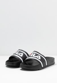 Zwarte sliders met een brede, gestructureerde band met witte accenten en het rode FILA-logo. Platte zool zonder hak en een gladde afwerking.