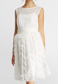 Robe blanche dotée d'un corsage ajusté avec superposition transparente et d'une jupe évasée ornée de détails texturés ressemblant à des plumes.