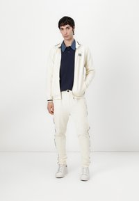 Cremefarbener Strick-Trainingsanzug mit dunklen Akzenten, Reißverschlussjacke, gerippten Bündchen und passenden Hosen. Das Modell trägt ein navyblaues Polohemd darunter.