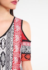 Top de hombros descubiertos con patrones en negro, blanco y rojo, que presentan diseños florales y geométricos, terminado con un contraste de ribete negro.