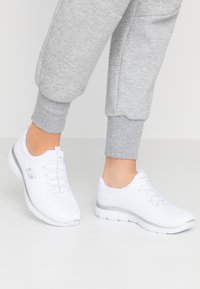 Scarpe sportive bianche con tomaia in rete, punta rotonda e un accento grigio sulla suola. Presentano lacci e logo sul lato.