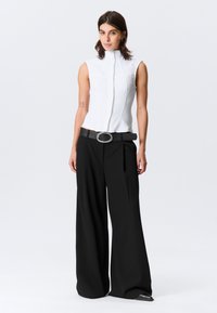 Haut blanc sans manches avec un col montant et un tissu texturé, associé à un pantalon noir à jambes larges et une ceinture noire avec une boucle argentée.