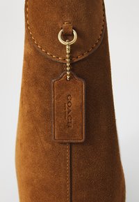 Brun ruskind håndtaske med en guldtonet lukning og et rektangulært mærke, der viser "COACH NEW YORK". Overfladen har en blød tekstur.