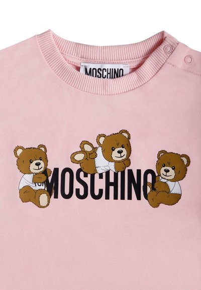 Różowa odzież z trzema brązowymi misiami w białych koszulach otaczającymi czarne logo marki "MOSCHINO".
