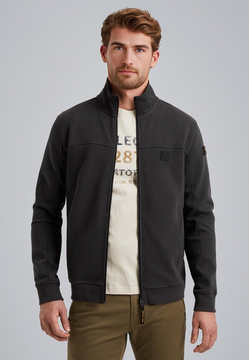 Veste zippée noire avec panneau supérieur texturé, section inférieure lisse, col montant et marquage subtil sur la poitrine et la manche.