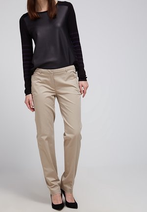Kvinde iført beige bukser med lige ben, sort bluse med lange ærmer og sorte højhælede sko, står foran en ensfarvet baggrund.